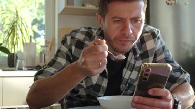 Ekose gömlekli bir adam akıllı telefon kullanırken beyaz kaseden yulaf lapası yiyor. Erkek kahvaltıda telefona bakıyor. Sosyal medya bağımlılığı, dikkat dağıtma ve dijital bağımlılık kavramı