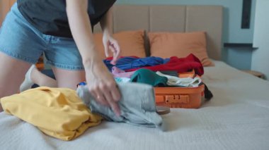 Tatile hazırlanan bir kadın, parlak turuncu bavul, mayo ve yaz kıyafetlerini yatağa koyuyor. Tatil hazırlığı