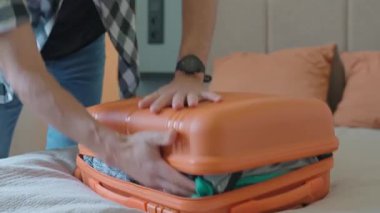 Yolculuktan önce turuncu bavulu kapatmaya çalışan bir adam. Erkek gezgin bir sürü giysi hazırlarken, seyahat için hazırlanırken zorlanır.