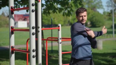 Spor giyimli bir adam yeşilliklerle çevrili açık hava spor salonunda esniyor. Atletik adam antrenmandan önce parkta ısınıyor. Zindelik, sağlıklı yaşam tarzı ve sağlık kavramı