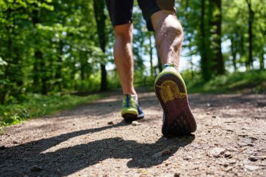 Güneşli orman yolunda yürüyen spor ayakkabıların alçak açılı görüntüsü. Güneşli bir günde parkta koşan erkek sporcuların yakın çekimi. Açık hava koşusu, fitness ve sağlıklı yaşam tarzı kavramı