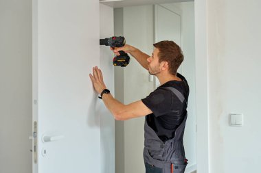 Adam kablosuz matkabı tutuyor ve modern iç mekanda beyaz kapı kolu takıyor. Tornavida kullanarak evin kapılarını tamir eden tamirci. Yenileme sırasında donanım kurulumu kavramı