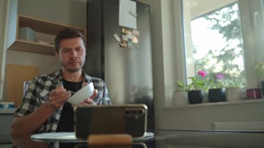 Ekose gömlekli bir adam akıllı telefon kullanırken beyaz kaseden yulaf lapası yiyor. Erkek kahvaltıda telefona bakıyor. Sosyal medya bağımlılığı, dikkat dağıtma ve dijital bağımlılık kavramı