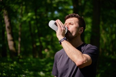 Ormanda koşarken siyah spor giyimli bir adam su içiyor. Aktif erkek sporcu Green Park 'ta koşarken su ile ferahlatıyor. Açık hava sporları ve sağlıklı yaşam tarzı kavramı