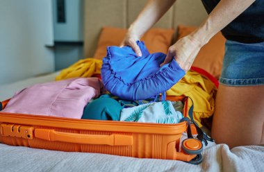 Kadın renkli kıyafetleri yatak odasında turuncu bavula katlıyor. Tatil hazırlığı, seyahat gereksinimleri ve paketleme süreci kavramı