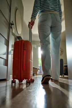 Dolaşan bavullarla kapıya doğru yürüyen gezginin alçak açılı görüntüsü. Koridorda turuncu bavulla yürüyen kişi. Ayrılma ve tatil için evden ayrılma kavramı
