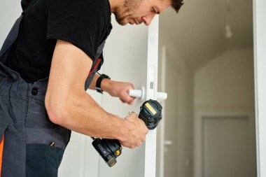 Adam kablosuz matkabı tutuyor ve modern iç mekanda beyaz kapı kolu takıyor. Tornavida kullanarak evin kapılarını tamir eden tamirci. Yenileme sırasında donanım kurulumu kavramı