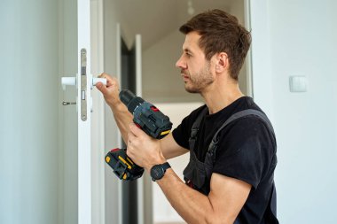 Adam kablosuz matkabı tutuyor ve modern iç mekanda beyaz kapı kolu takıyor. Tornavida kullanarak evin kapılarını tamir eden tamirci. Yenileme sırasında donanım kurulumu kavramı