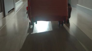 Dolaşan bavullarla kapıya doğru yürüyen gezginin alçak açılı görüntüsü. Koridorda turuncu bavulla yürüyen kişi. Ayrılma ve tatil için evden ayrılma kavramı