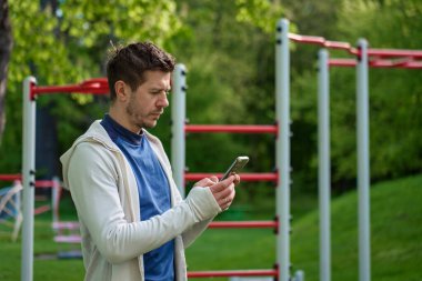 Spor giyim bölümündeki adam antreman istasyonundaki telefonu kontrol ediyor. Spor kıyafetleri içinde sporcu akıllı telefonuyla egzersiz barlarının yanında duruyor.