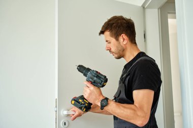 Adam kablosuz matkabı tutuyor ve modern iç mekanda beyaz kapı kolu takıyor. Tornavida kullanarak evin kapılarını tamir eden tamirci. Yenileme sırasında donanım kurulumu kavramı