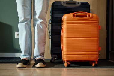 Kot pantolon ve ayakkabı giyen, bavulun yanında duran seyahat için evden çıkmaya hazır bir kadın. Ayrılmadan önce kapının yanında kadın ayağı ve bavul var.