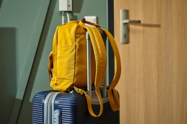 Kapıda duran koyu renkli bavul ve sarı sırt çantası. Kalkışa hazırız. Yolculuk ya da seyahat hazırlığı