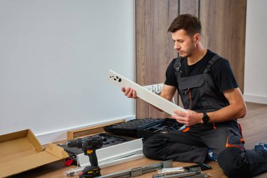 Tulumlu adam mobilyaları birleştiriyor, beyaz paneli inceliyor ve aletlerle çevrili. İş kıyafetleri içinde tamirci parçaları montaja hazırlıyor. DIY mobilya projesi