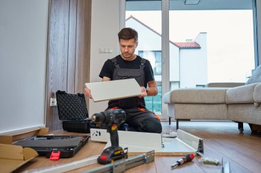 Tulumlu adam mobilyaları birleştiriyor, beyaz paneli inceliyor ve aletlerle çevrili. İş kıyafetleri içinde tamirci parçaları montaja hazırlıyor. DIY mobilya projesi