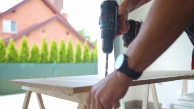 İş önlüklü bir adam kablosuz matkap kullanarak lamine ahşap tahtada delik açıyor. Erkek marangoz atölyede çalışıyor, tezgahta çipleri deliyor.