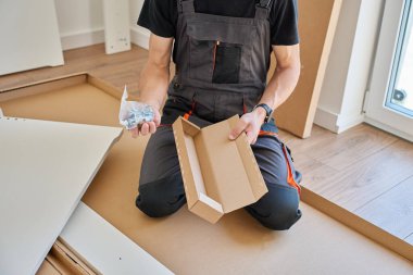 İş tulumlu bir adam, kutulu mobilya panelleri ve aletlerin yanında diz çökerken vida ve teçhizatla plastik torba açar. Mobilya kurulumu