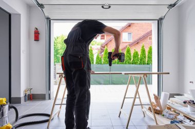 İşçi, evdeki garaj atölyesindeki testere atlarıyla desteklenen tahta tahtada matkap kullanıyor. Adam atölyede çalışıyor, mobilya montajı için yonga tahtasına delik açıyor.