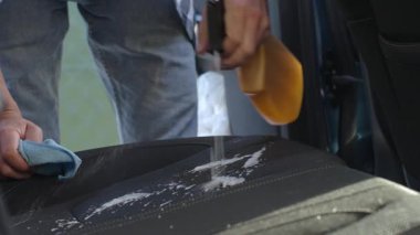 Kişi, oto koltuğuna temizlik köpüğü püskürtüp mikrofiber bez ile siler. İç temizlik sırasında. Araç döşemesi bakımı