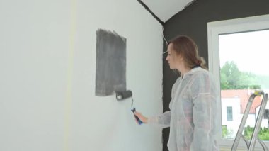 Koruyucu takım elbiseli, beyaz pantolonlu, siyah baskılı, yenileme sırasında tekerlekli paten kullanan bir kadın. İç duvarı boyayan kadın işçi. Evde tadilat ve iyileştirme kavramı