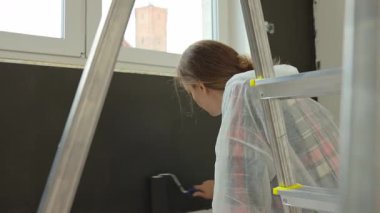 Koruyucu takım elbiseli kadın merdivenin üzerinde duruyor ve duvarları siyaha boyuyor. Ev yapımı sırasında iç boyama işlemi