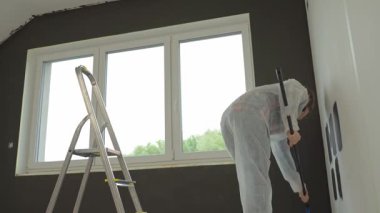 Koruyucu takım elbiseli, beyaz pantolonlu, siyah baskılı, yenileme sırasında tekerlekli paten kullanan bir kadın. İç duvarı boyayan kadın işçi. Evde tadilat ve iyileştirme kavramı