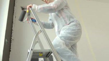 Koruyucu giysili kadın elinde boya silindiriyle merdiveni tırmanıyor ve siyah aksan duvarını boyamaya hazırlanıyor. Modern apartman içinde ev yenileme