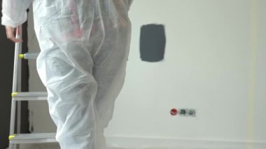 Koruyucu giysili kadın elinde boya silindiriyle merdiveni tırmanıyor ve siyah aksan duvarını boyamaya hazırlanıyor. Modern apartman içinde ev yenileme