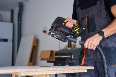 İş elbisesi işçisi elektrikli yapboza elektrikli hortum bağlıyor atölyedeki tezgahta marangozluk yapmak için