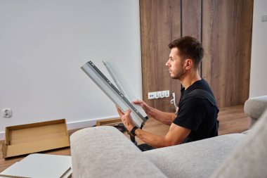 Tulumlu adam mobilyaları birleştiriyor, beyaz paneli inceliyor ve aletlerle çevrili. İş kıyafetleri içinde tamirci parçaları montaja hazırlıyor. DIY mobilya projesi