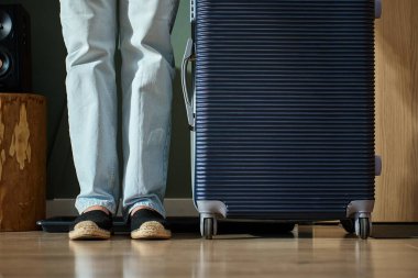 Kot pantolon ve ayakkabı giyen, bavulun yanında duran seyahat için evden çıkmaya hazır bir kadın. Ayrılmadan önce kapının yanında kadın ayağı ve bavul var.