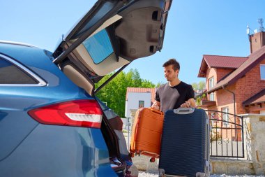 Banliyödeki tuğla evin önünde park edilmiş arabanın yanında turuncu ve mavi bavullar tutan adam. Bagajları araca yüklüyorum. Taşınma günü, taşınma ya da seyahat hazırlığı kavramı