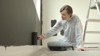 Koruyucu takım elbiseli bir kadın modern odada yenileme aşamasındaki fırçayla iç duvarı siyaha boyuyor.