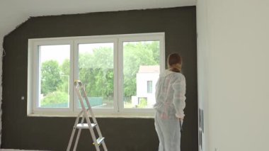 Koruyucu takım elbiseli, beyaz pantolonlu, siyah baskılı, yenileme sırasında tekerlekli paten kullanan bir kadın. İç duvarı boyayan kadın işçi. Evde tadilat ve iyileştirme kavramı