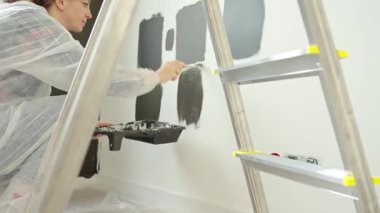 Koruyucu giysili kadın, fırça kullanarak beyaz duvara farklı koyu boya örnekleri uyguluyor. Modern evdeki iç yenileme projesi sırasında siyah ve koyu gri boya örneklerini test eden kadın ressam.