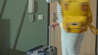 Kadın sarı sırt çantasını giyer, bavulunu alır ve apartmanın kapısından çıkar. Çanta ve bavulla evden çıkan kişi. Kot pantolon ve süveter giyen bayan gezgin seyahat ya da evden ayrılma hazırlıkları yapıyor.