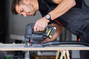 İş elbisesi işçisi garajda tahta kesmek için kablosuz elektrikli yapboz kullanıyor. Atölyede çalışan yetenekli bir marangoz. Ev tadilatı yapan adam.