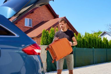 Güneşli bir günde turuncu bavulu bagaja yükleyen adam. Yaz seyahatine, tatile ya da bavulla yolculuğa hazırlanma kavramı