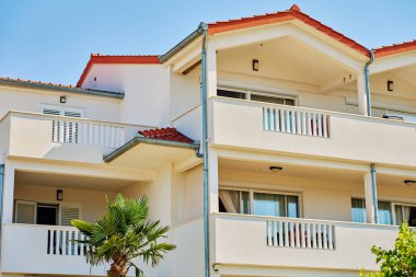 White residential building with balconies, red tiled roof and open windows under blue sky. Facade of modern villa with open terraces and palm tree. Mediterranean summer housing for vacations