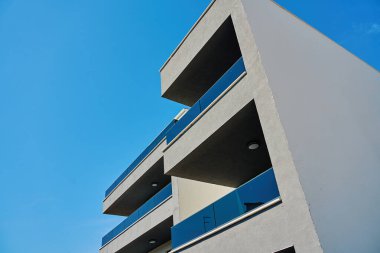 Mavi gökyüzünün altında minimalist balkonları ve cam parmaklıkları olan modern bir apartman. Çağdaş konut mimarisi, geometrik tasarım ve kentsel konut kavramı