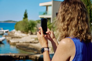 Güneşli bir günde mavi elbiseli bir kadının deniz kenarında akıllı telefonuyla fotoğraf çekişi. Kadın turist, deniz manzarasını telefonla çekiyor. Yaz tatili, seyahat ve teknoloji konsepti