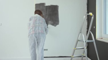 Koruyucu takım elbiseli, beyaz pantolonlu, siyah baskılı, yenileme sırasında tekerlekli paten kullanan bir kadın. İç duvarı boyayan kadın işçi. Evde tadilat ve iyileştirme kavramı
