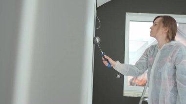 Koruyucu takım elbiseli, beyaz pantolonlu, siyah baskılı, yenileme sırasında tekerlekli paten kullanan bir kadın. İç duvarı boyayan kadın işçi. Evde tadilat ve iyileştirme kavramı