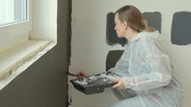 Kadın cam kenarındaki duvarı fırça ve boya tepsisiyle boyuyor, koruyucu takım elbise giyiyor. Ev yenileme, DIY dekorasyon ve duvar boyama kavramı
