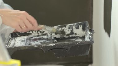 Close-up of hand using brush to apply dark paint to wall near paint tray. Female painter applying color swatches on white wall. Concept of interior renovation and wall painting