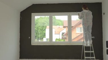 Koruyucu takım elbiseli kadın merdivenin üzerinde duruyor ve duvarları siyaha boyuyor. Ev yapımı sırasında iç boyama işlemi