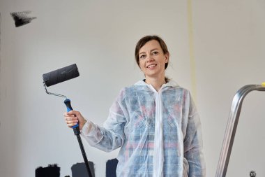 Koruyucu tulumlu gülümseyen kadın odada duvarları boyarken boya silindiri tutuyor. Ev yenileme ve iç yenileme kavramı