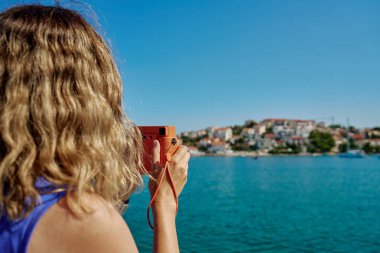 Denize yakın bir yerde elinde fotoğraf makinesi olan bir kadın. Sahildeki tatil köyünde arka planda bulanık şehir olan bir bayan turist. Seyahat ve yaz tatili konsepti