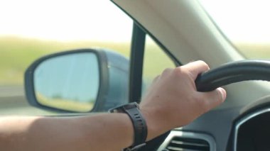 Arabanın direksiyonuna yakın plan dikiz aynasına yansıyan yol. Erkek sürücü araba kullanıyor, içeriden.
