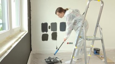 Koruyucu takım elbiseli, beyaz pantolonlu, siyah baskılı, yenileme sırasında tekerlekli paten kullanan bir kadın. İç duvarı boyayan kadın işçi. Evde tadilat ve iyileştirme kavramı